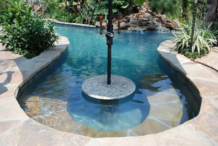 an umbrella is in the middle of a pool with rocks and plants around it,