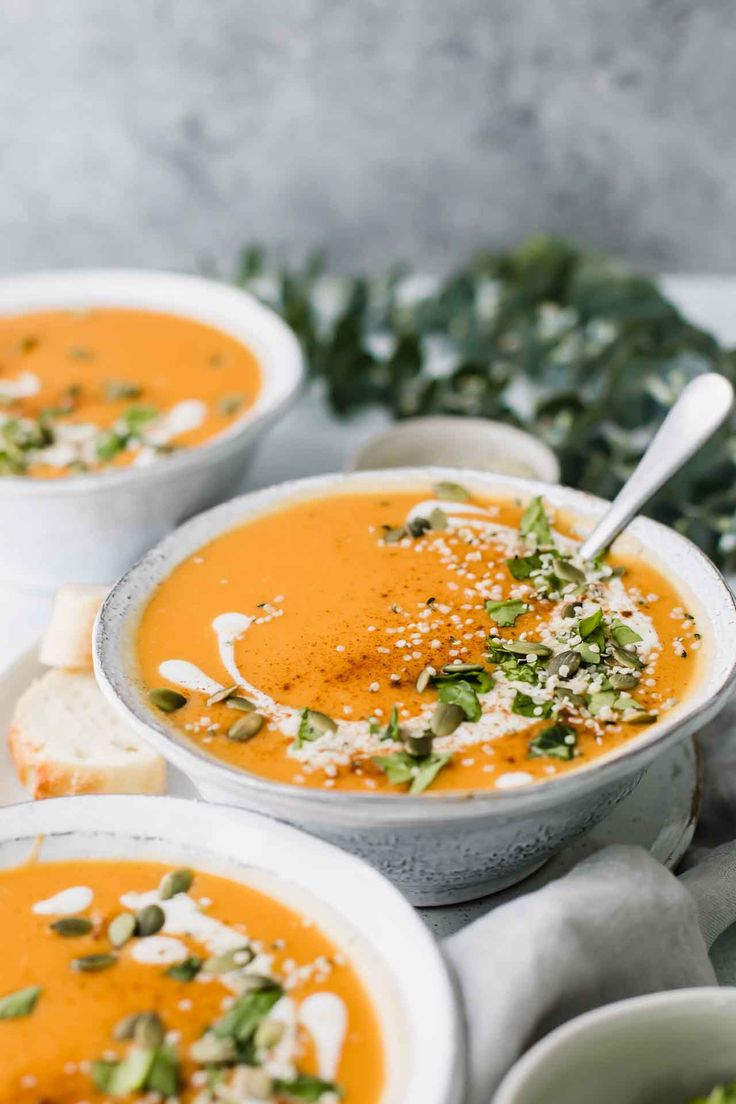 three bowls of carrot soup with garnishes and bread on the side, ready to be eaten