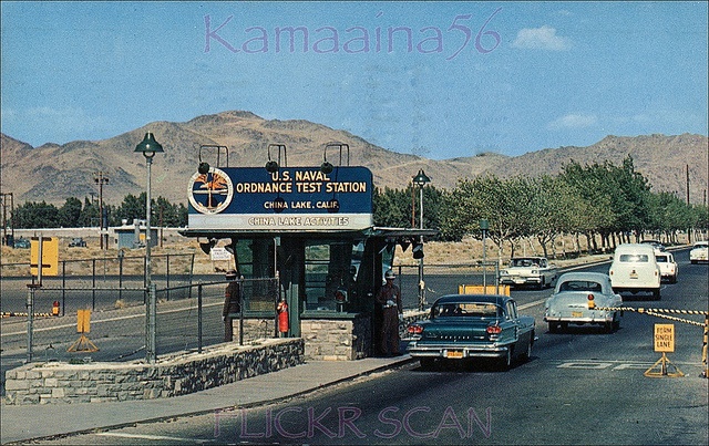 cars are driving down the road in front of a fenced off parking lot with mountains in the background