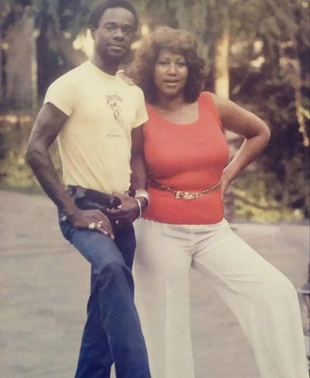 a man and woman standing next to each other in front of some trees with their hands on their hipss