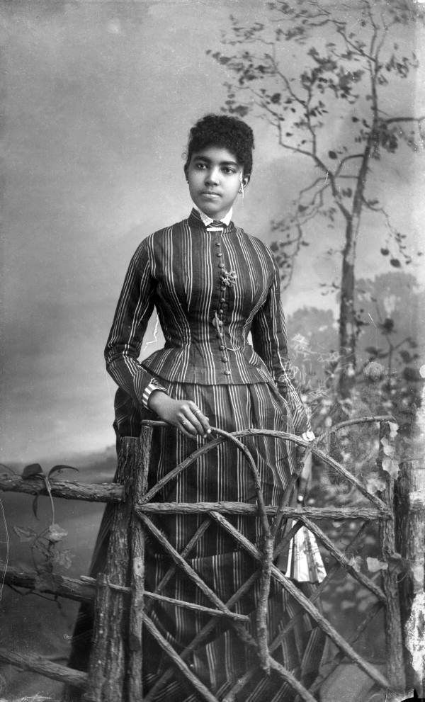 an old black and white photo of a woman in a striped dress standing on a fence