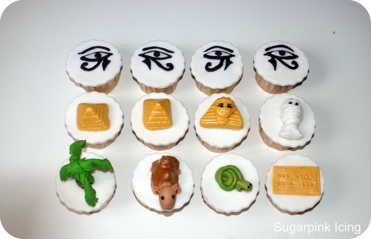 cupcakes decorated with egyptian symbols are displayed on a white table top, including an eye and other items