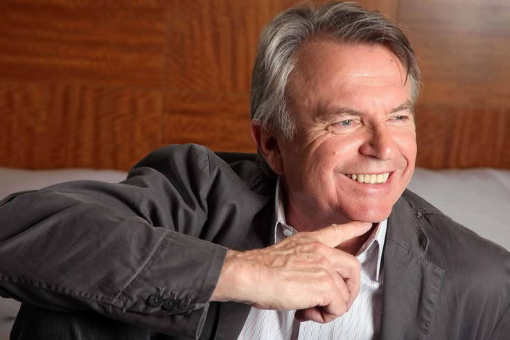 an older man smiles while sitting on a bed with his hand under his chin and looking at the camera