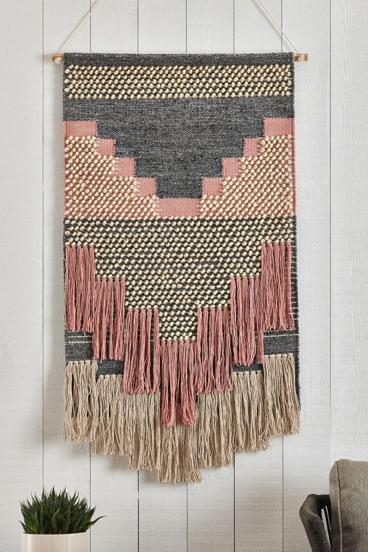 a woven wall hanging with fringes on it next to a chair and potted plant