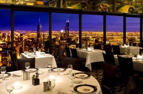 a dining room with tables and chairs overlooking the city at night in front of large panoramic windows