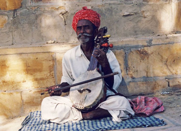 an old man sitting on the ground with a musical instrument in his hand and wearing a red turban