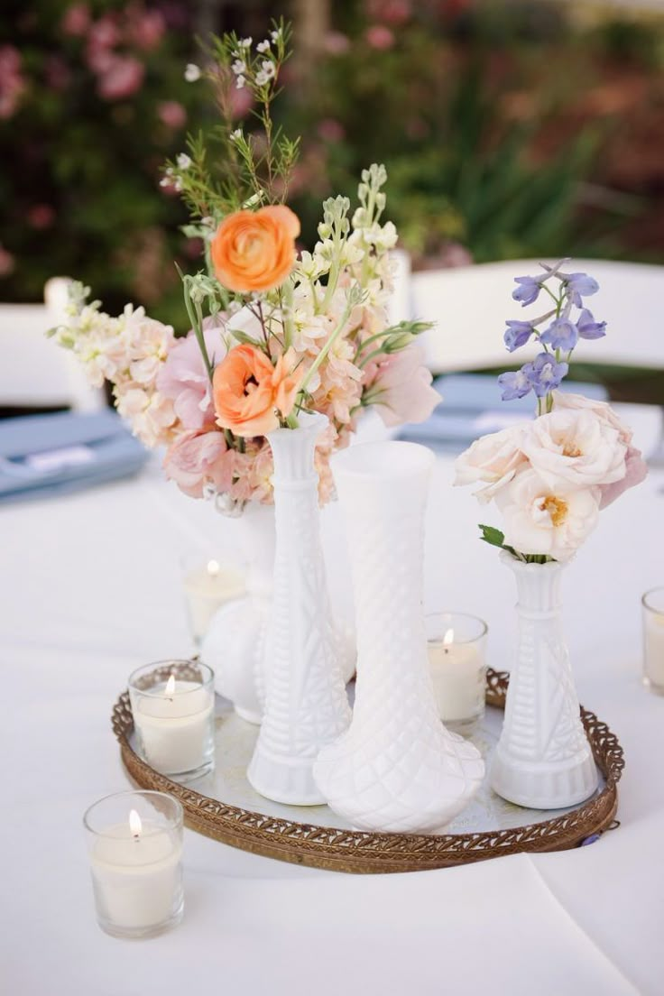 three white vases filled with flowers sitting on top of a table next to candles