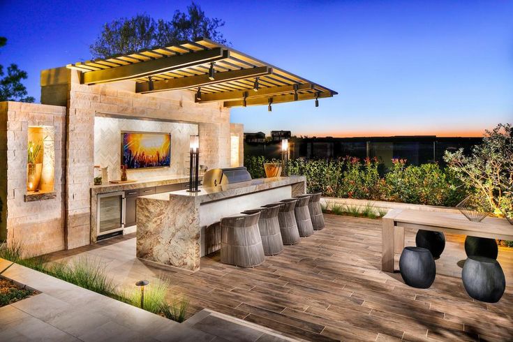 an outdoor kitchen with grill and seating area at dusk, surrounded by trees and shrubs