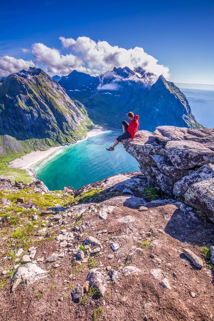 a man sitting on the edge of a cliff looking out over water and mountains in norway