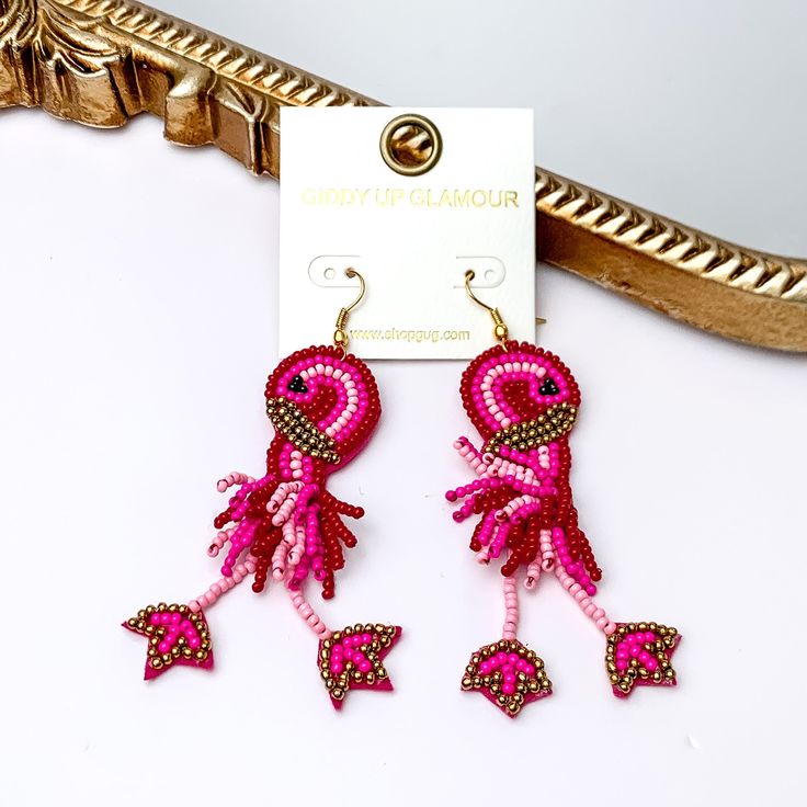 These earrings include multiple color pink beads and a gold detailing. Hanging from the bottom of the flamingo shaped beaded pendant has dangling legs. These earrings are pictured leaning agasint a gold mirror on a white background. Trendy Beaded Earrings For The Beach, Trendy Dangling Beads Earrings For Beach, Trendy Beach Earrings With Dangling Beads, Pink Jewelry For Beach Party, Bohemian Pink Jewelry For Beach Party, Adjustable Beaded Earrings With Dangling Beads For Beach, Pink Jewelry For Beach Party Season, Bohemian Beaded Dangling Earrings For Vacation, Summer Party Beaded Drop Earrings