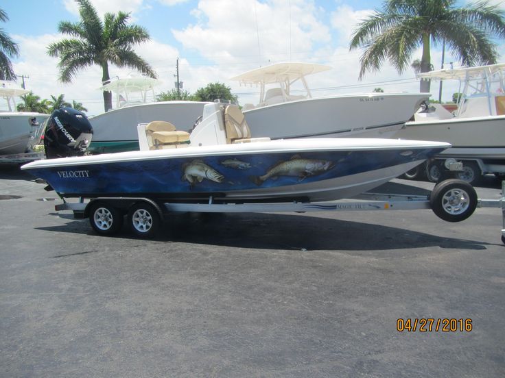 a blue and white boat parked in a parking lot next to other boats with palm trees behind it