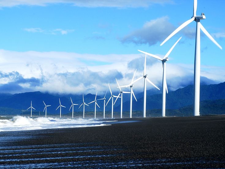 several wind turbines on the beach with mountains in the background and clouds in the sky