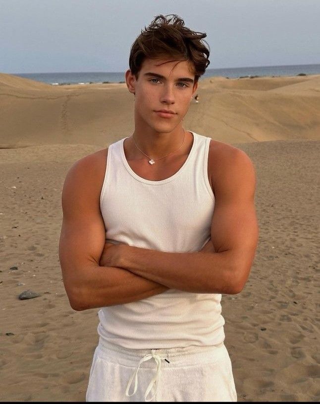 a man in white shirt standing on beach next to the ocean with his arms crossed