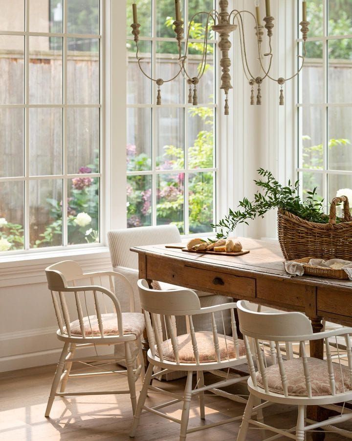 a dining room table with four chairs and a basket on top of it, in front of two windows