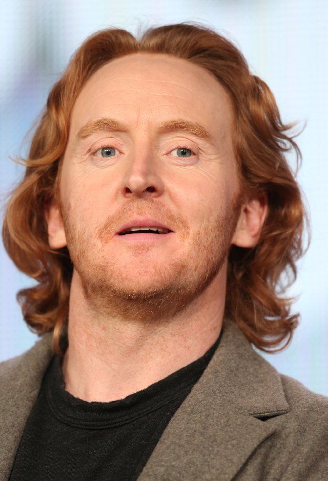 a man with long red hair wearing a gray suit and black t - shirt is looking at the camera