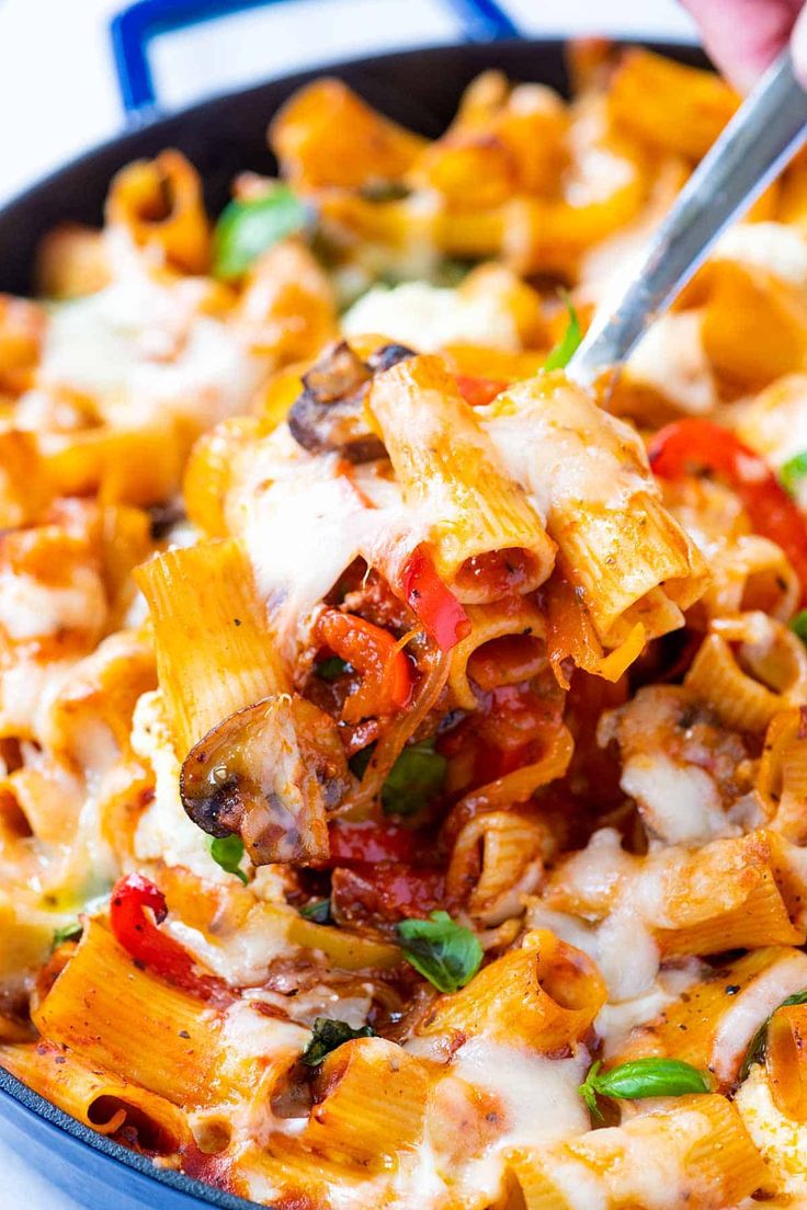 a hand holding a fork full of pasta with sauce, cheese and vegetables in a skillet