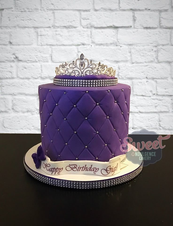 a purple birthday cake with a tiara on top is sitting in front of a white brick wall