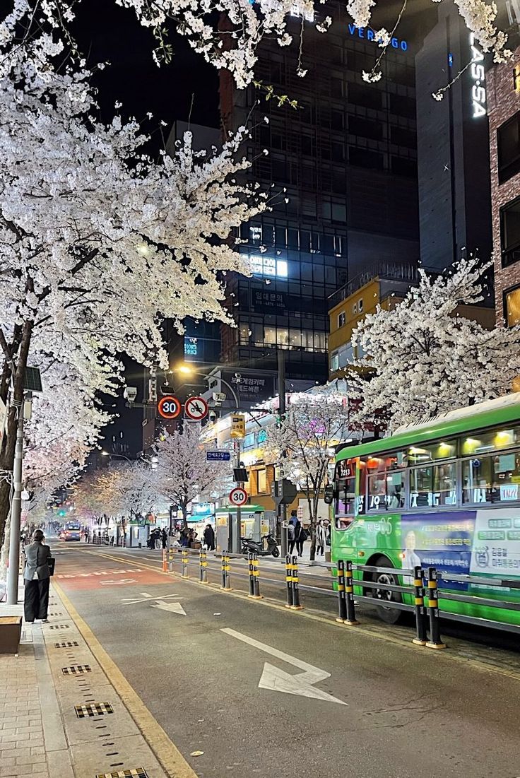 a green bus driving down a street next to tall buildings and trees with white flowers