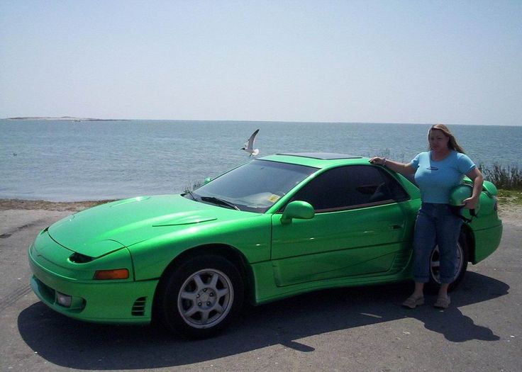 a woman standing next to a green sports car near the ocean with a seagull perched on top