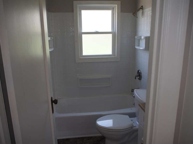 a white toilet sitting next to a bath tub in a bathroom under a window on top of a tiled floor