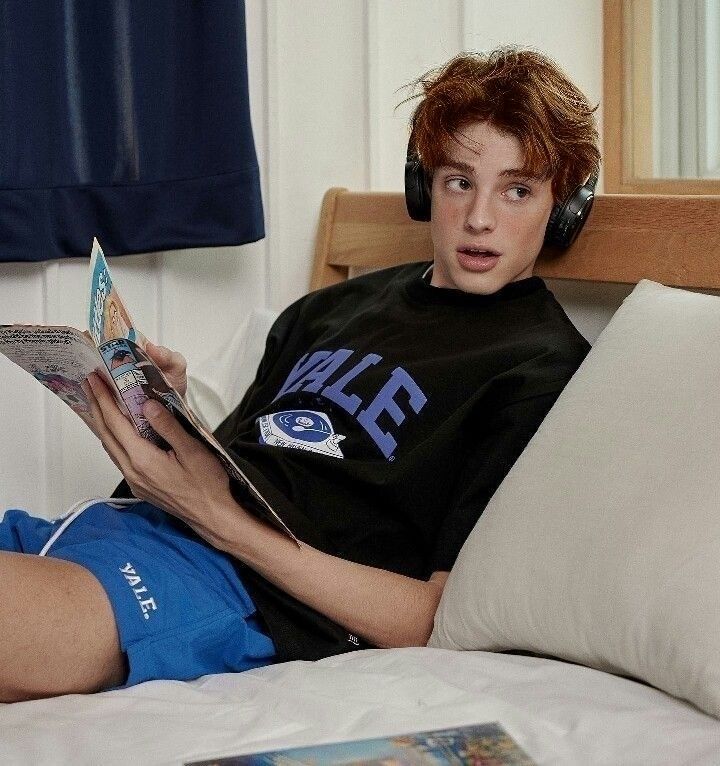 a boy laying in bed with headphones on reading a book