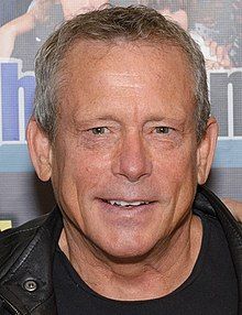 a close up of a person wearing a black shirt and leather jacket with a poster in the background