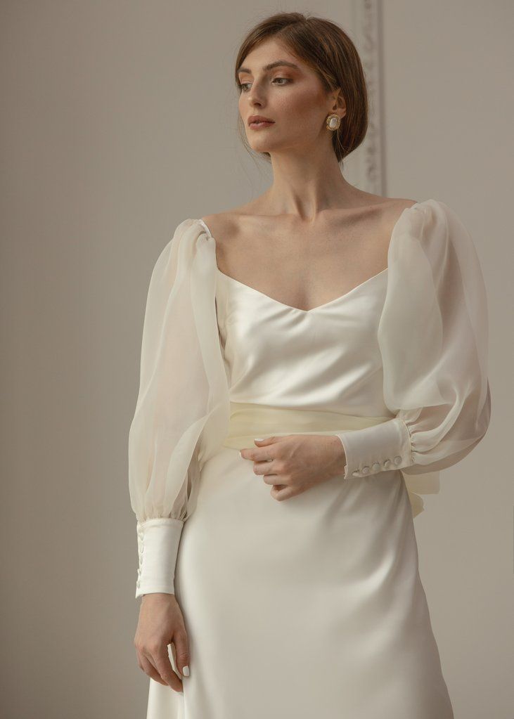 a woman wearing a white dress with long sleeves and pearls on the neck, standing in front of a mirror