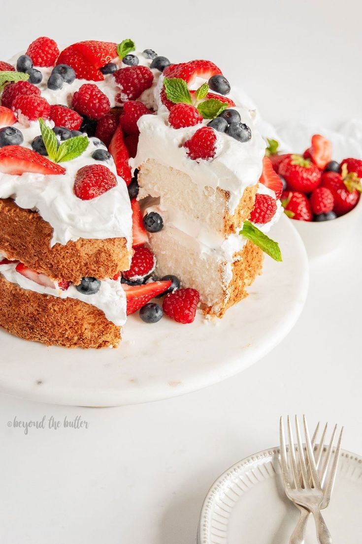 a white plate topped with a cake covered in whipped cream and berries next to a bowl of strawberries