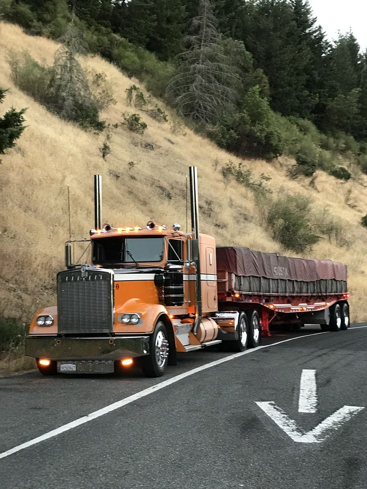 an orange semi truck driving down the road next to a hill with trees on it