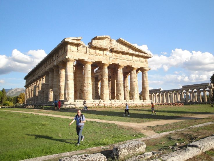 people are walking around in front of an ancient building with columns on it's sides