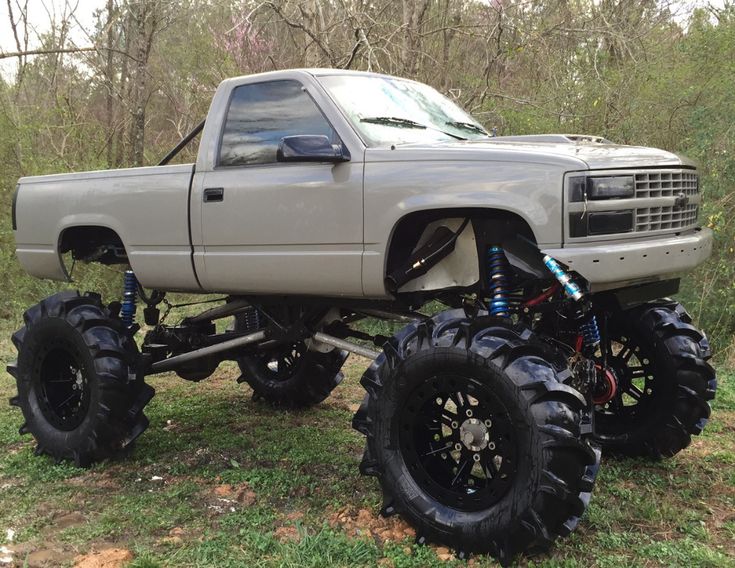 a silver truck with large tires parked in the grass next to some trees and bushes