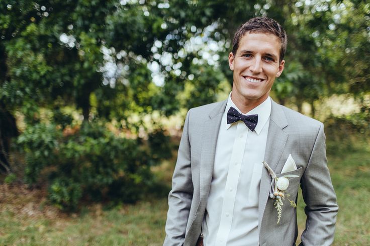 a man in a gray suit and bow tie smiles at the camera while wearing a boutonniere