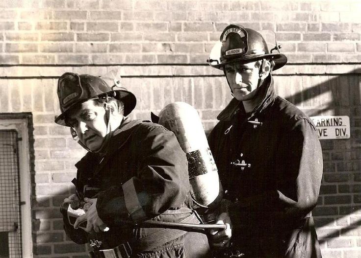 two firemen standing next to each other in front of a brick wall and building