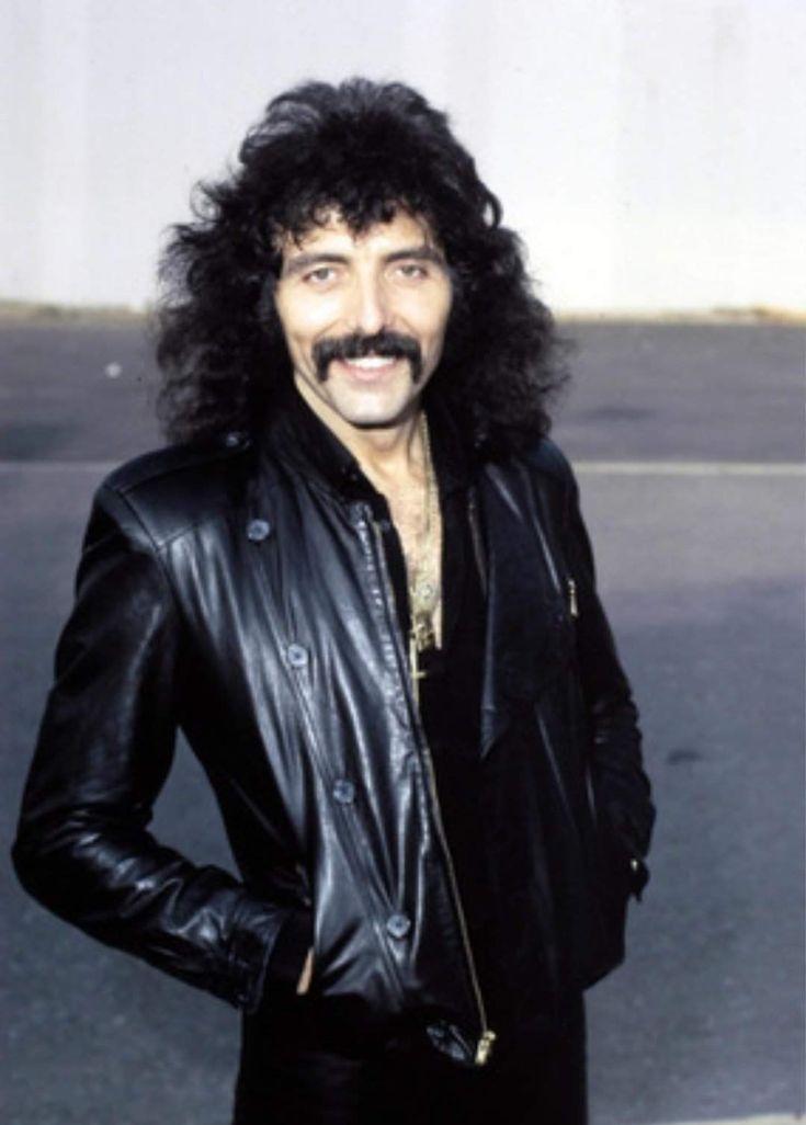 a man with long hair and a moustache wearing a black leather jacket standing in front of a building