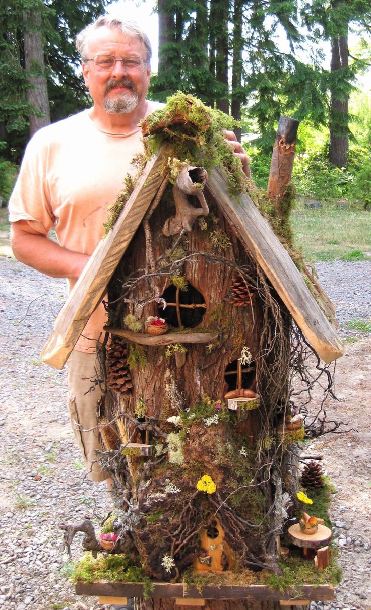 a man standing next to a tree house made out of wood and mossy branches