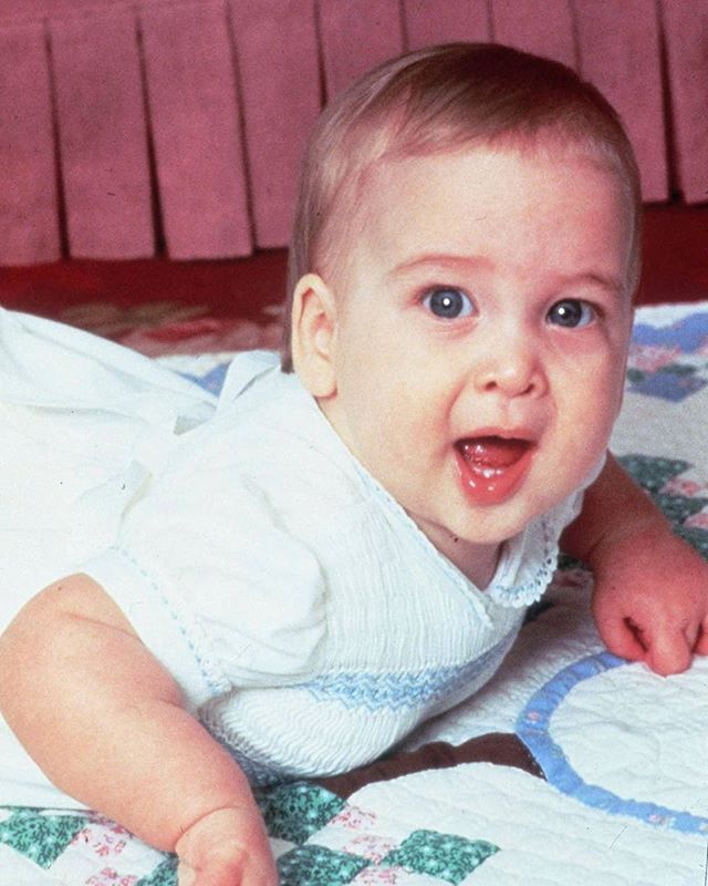 a baby laying on top of a bed wearing a white shirt and diaper with his tongue out