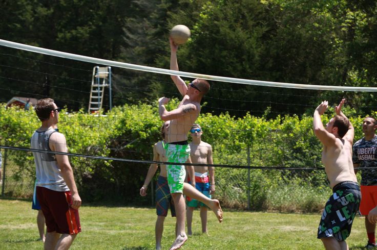 men playing volleyball on the grass in front of some bushes and trees, with one man reaching up to hit the ball