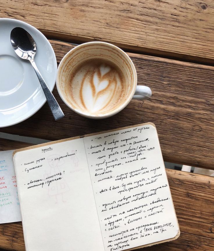 a cup of coffee on top of a wooden table next to an open book and spoon