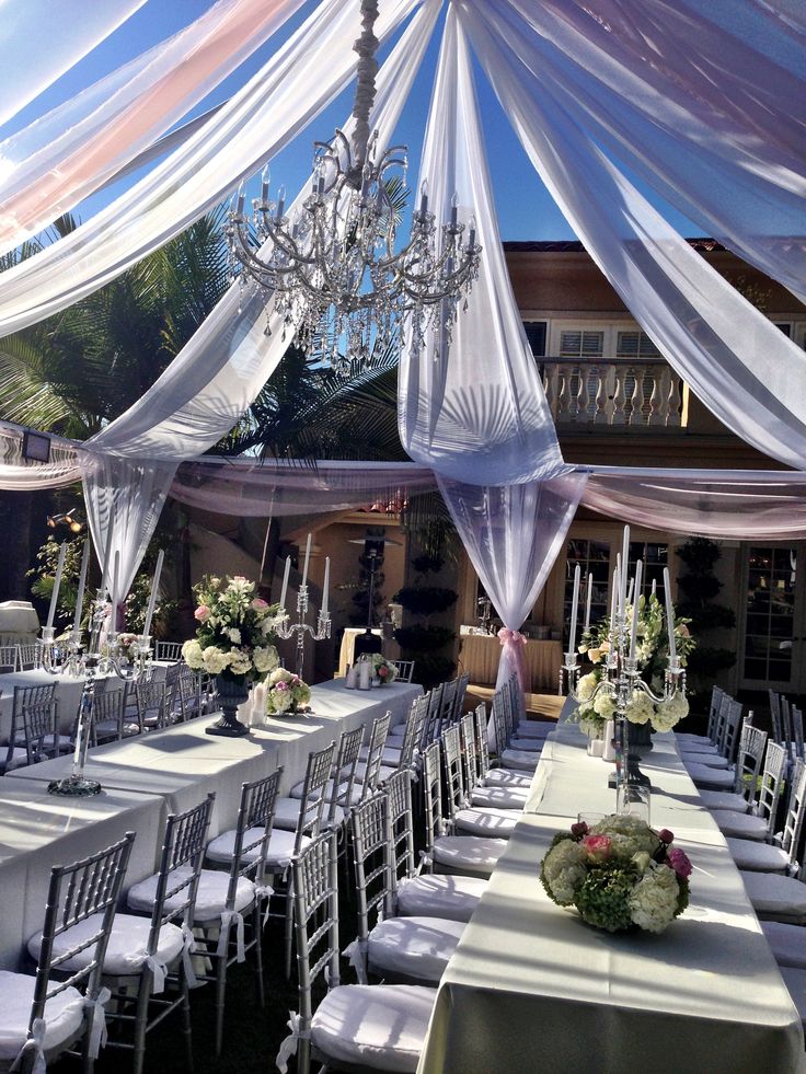 an outdoor wedding setup with white chairs and chandelier hanging from the ceiling over tables