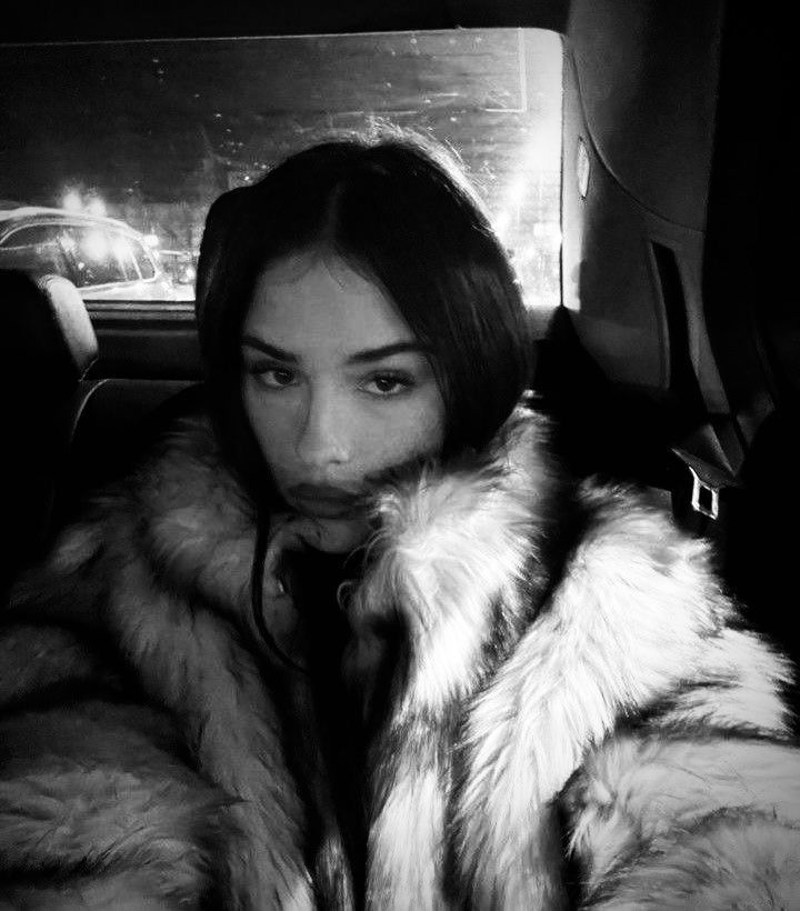 a woman wearing a fur coat in the back seat of a car, looking at the camera