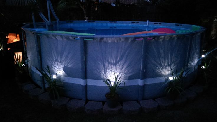 an above ground swimming pool lit up at night with plants and lights around the edge