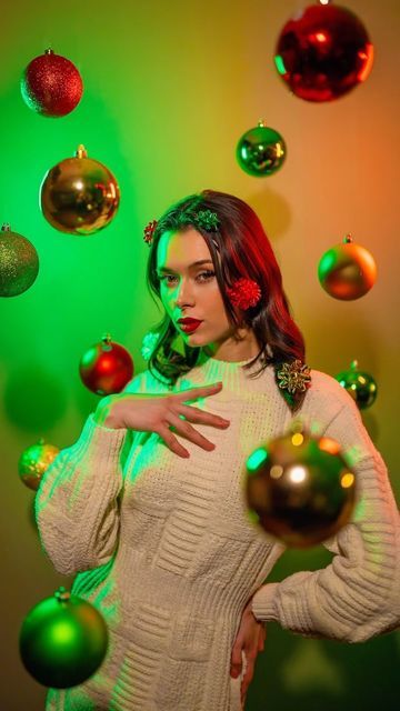 a woman in a white sweater surrounded by green and red christmas ornaments, with her hands on her hips