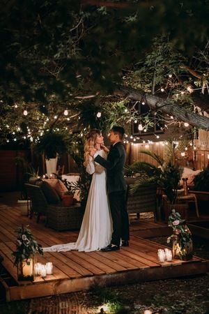 a bride and groom standing on a wooden deck surrounded by lit up trees at night