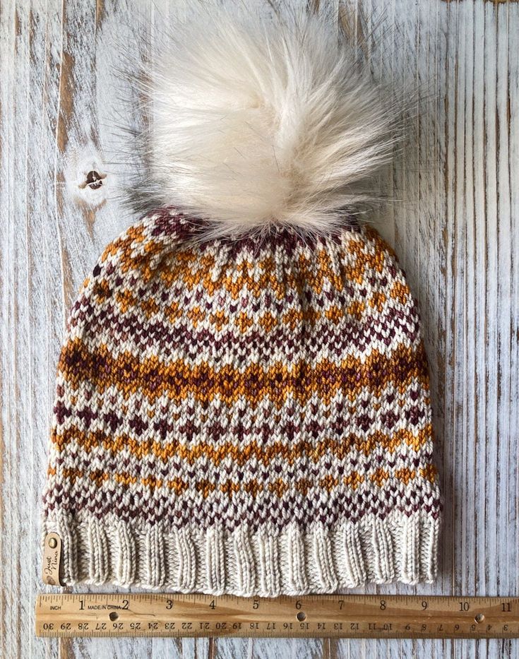 a knitted hat with a white pom - pom sitting on top of a ruler