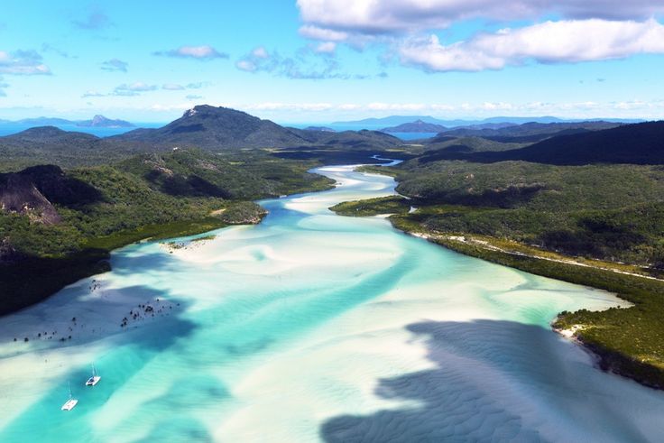 an aerial view of white sand and blue water in the middle of green land with mountains