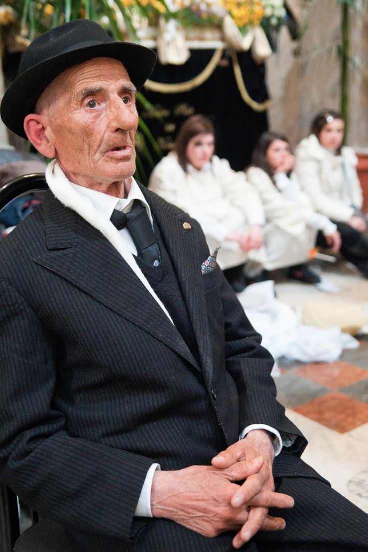 an older man in a suit and hat sitting on a chair with other people behind him