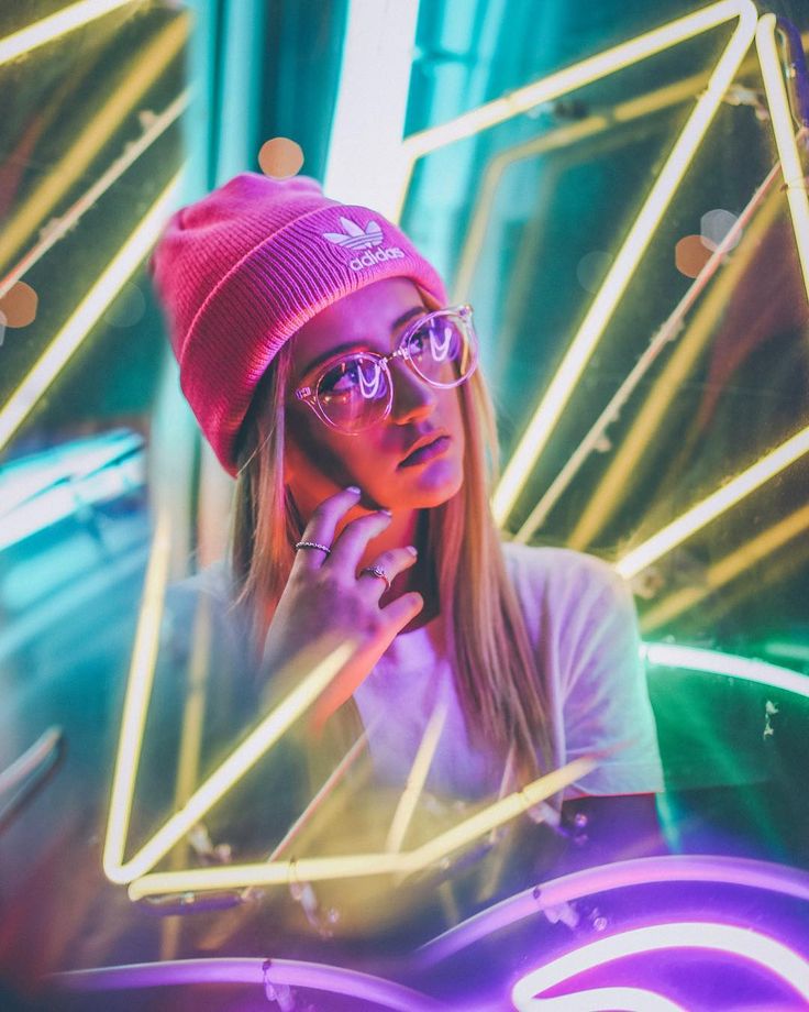 a woman wearing glasses and a beanie talks on her cell phone in front of neon lights