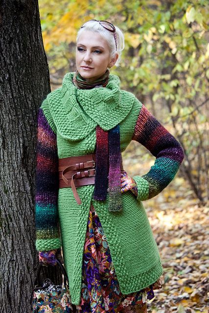 a woman standing in front of a tree wearing a green coat and colorful scarfs