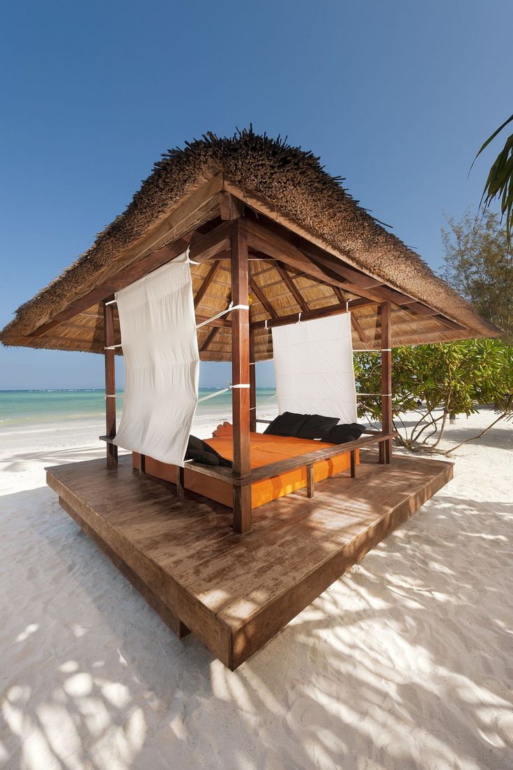 a bed sitting on top of a sandy beach under a thatch roofed roof next to the ocean