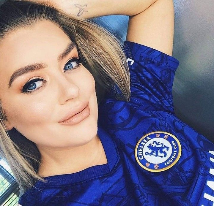 a woman with blonde hair and blue eyes wearing a chelsea shirt looking at the camera